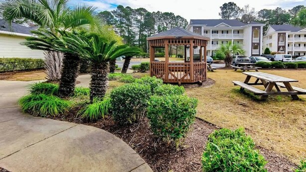 A gazebo in a landscaped garden area with a picnic table nearby, surrounded by well-maintained shrubs and palm trees. - River Oaks Unit 25A Condo (Myrtle Beach)