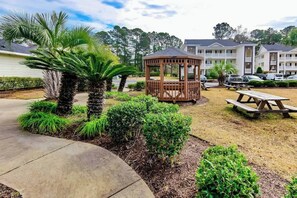 A gazebo in a landscaped garden area with a picnic table nearby, surrounded by well-maintained shrubs and palm trees. - River Oaks Unit 25A Condo (Myrtle Beach)