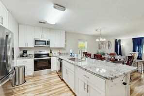 A modern kitchen with white cabinetry, a granite countertop, stainless steel appliances, and a dining area with red chairs. - River Oaks Unit 25A Condo (Myrtle Beach)