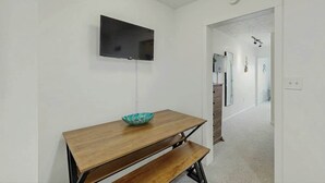 Um quarto com uma mesa de madeira, uma tigela, uma televisão de tela plana na parede e um corredor que dá para outro quarto.