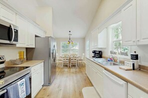 A kitchen with white cabinets, a stainless steel refrigerator, a microwave, and a dining area with a table and chairs. - Pawleys Beach Retreat (Pawleys Island)