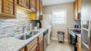 A kitchen with wooden cabinets, granite countertops, a built-in dishwasher, and a window with blinds. - Whalecome Inn Retreat (Myrtle Beach)