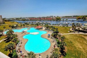 Ein großer Außenpool mit verschiedenen Bereichen, umgeben von Palmen und Liegestühlen, mit einem Hafen und Gebäuden im Hintergrund.
