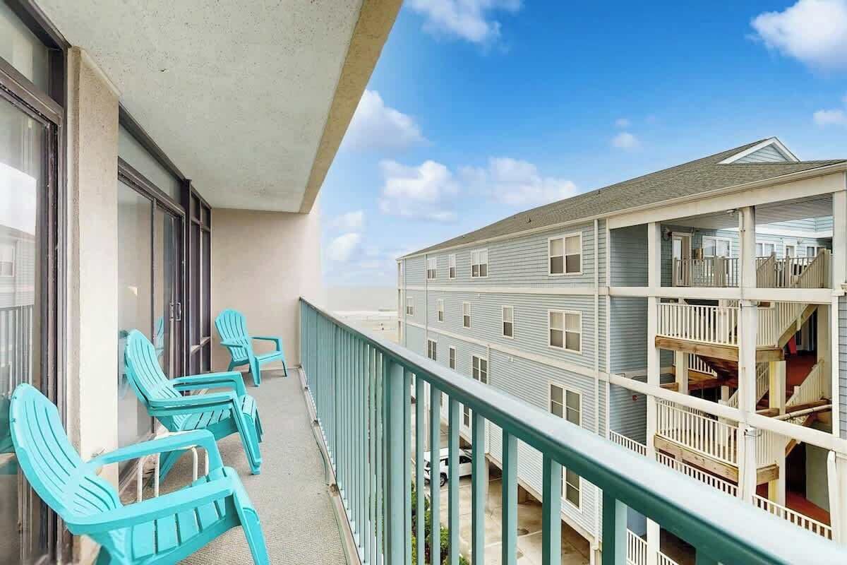 Ein Balkon mit türkisfarbenen Stühlen, Blick auf ein mehrstöckiges Gebäude und einen klaren Himmel.