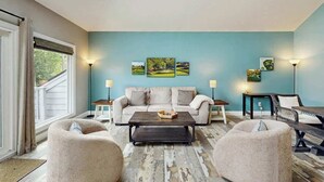 Ein Wohnzimmer mit einer Couch, zwei Sesseln, einem Couchtisch und einem Esstisch. Drei Wandlampen und ein Gemälde befinden sich über der Couch.