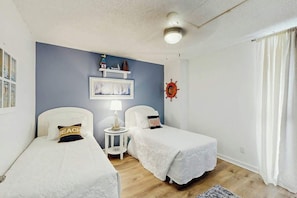 A bedroom with two single beds, a small table with a lamp, a wall-mounted shelf with decorative items, and a ship's wheel decoration. - Brigadune Unit 12B Condo (Myrtle Beach)