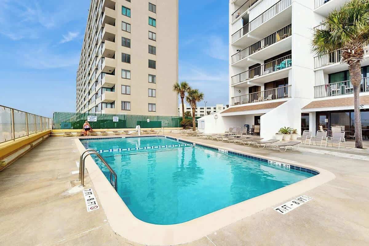 A swimming pool with a clear blue water, surrounded by lounge chairs and a palm tree, with a tall building in the background.