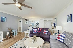 Ein Wohnzimmer mit Deckenventilator, einem runden Couchtisch mit Dekoration, einem violetten Sofa mit nautisch gestalteten Kissen und einem grauen Sofa mit gestreiften Kissen.