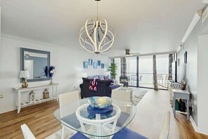 A modern living room with a glass dining table, a unique chandelier, and a balcony with a view. - Brigadune Unit 12B Condo (Myrtle Beach)