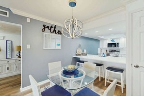 A modern dining area with a glass table and white chairs, a decorative ceiling light, and a kitchen in the background. - Brigadune Unit 12B Condo (Myrtle Beach)