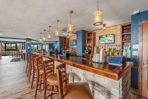 A bar area with a wooden counter, multiple bar stools, hanging pendant lights, and a television screen. - The Patricia Grand 1804 (Myrtle Beach)
