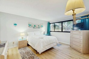 A bedroom with a bed, bedside tables, a dresser, a lamp, artwork on the wall, and a window with curtains. - Holiday Towers Unit 205 (Myrtle Beach)