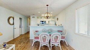 A dining area with a glass-top table and white chairs, a kitchen in the background, and a chandelier. - Carolina Yacht Landing E-13 (Little River)