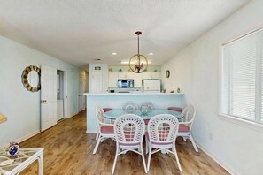 A dining area with a glass-top table and white chairs, a kitchen in the background, and a chandelier. - Carolina Yacht Landing E-13 (Little River)