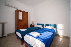 Quarto de hotel com duas camas, um guarda-roupa de madeira, uma mesa de cabeceira com abajur e um radiador.