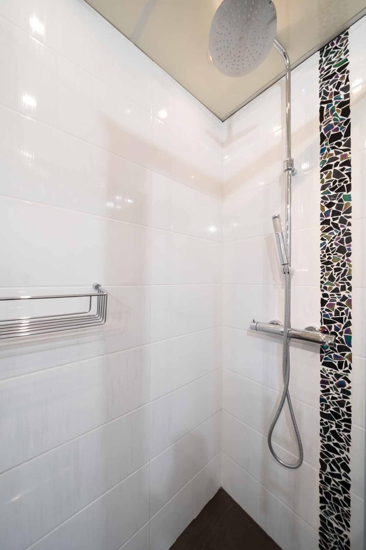 A modern shower with a rain showerhead and a handheld showerhead, a chrome towel rack, and a mosaic tile accent on the wall.