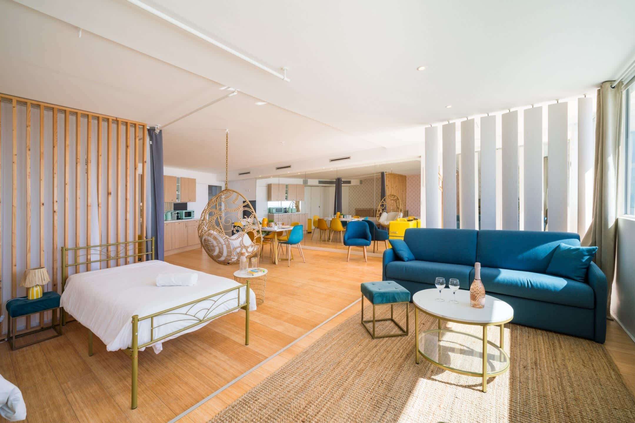 Een moderne hotelkamer met een bed, een blauwe bank, een zithoek en een keuken op de achtergrond.
