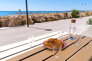 Une table avec une assiette de croissants, une bouteille d’eau et deux verres, le tout placé en plein soleil, avec vue sur une côte rocheuse et un phare visible au loin.