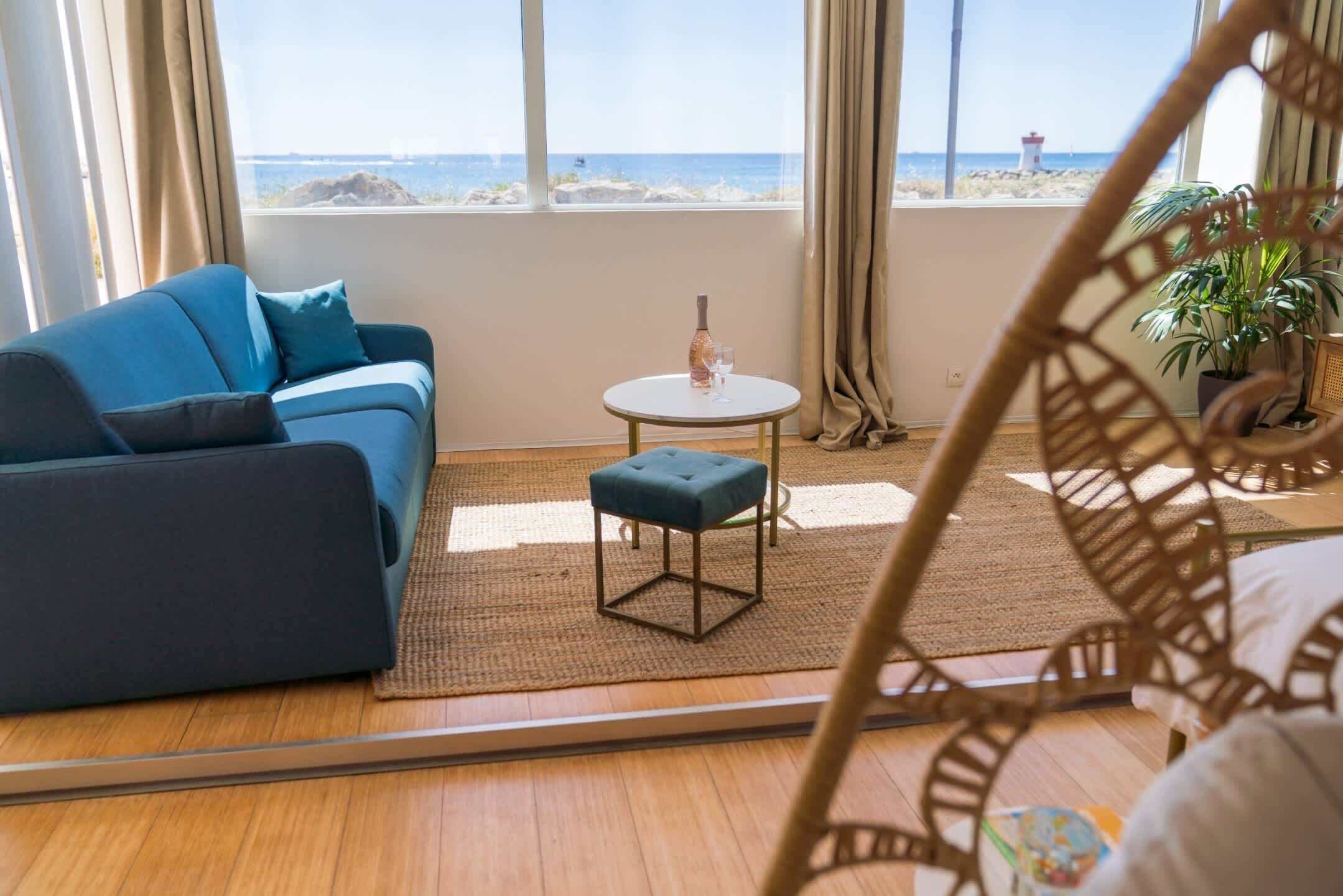 Ein Zimmer mit einem blauen Sofa, einem kleinen runden Tisch mit einer Flasche und einem Glas, Holzdielen und Meerblick durch große Fenster.
