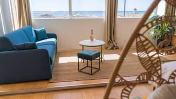 Ein Zimmer mit einem blauen Sofa, einem kleinen runden Tisch mit einer Flasche und einem Glas, Holzdielen und Meerblick durch große Fenster.