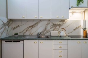 A kitchen with marble backsplash, white cabinets with gold handles, a built-in dishwasher, and a kettle on the counter. - Parthenopi suite – proche palais et ville (Cannes)