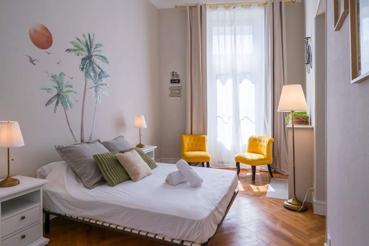 A hotel room with a bed, two chairs, a nightstand, a lamp, a wall with a palm tree mural, and a window with curtains.