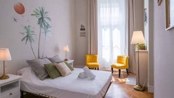 Ein Hotelzimmer mit einem Bett, zwei Stühlen, einem Nachttisch, einer Lampe, einer Wand mit Palmenmotiv und einem fenster mit Vorhängen.
