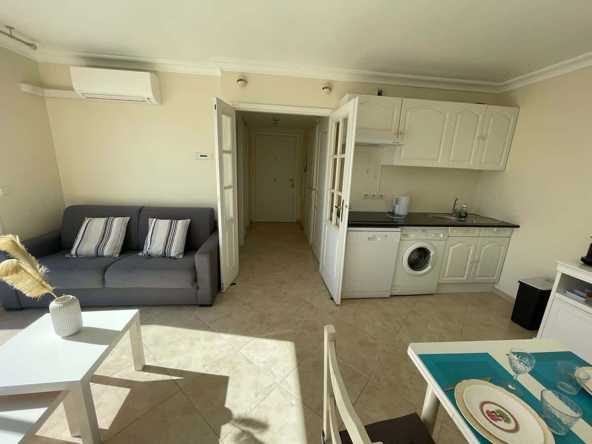 Un espacio habitable compacto con sofá, mesa de comedor, zona de cocina con lavadora y una pequeña mesa con un objeto decorativo.
