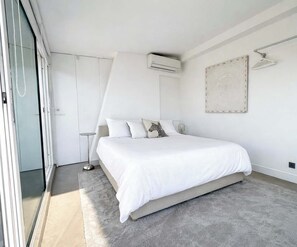 A modern bedroom with a large bed, a bedside table, an air conditioning unit, and a sliding glass door. - Riviera rooftop - front de mer avec terrasse (Antibes)