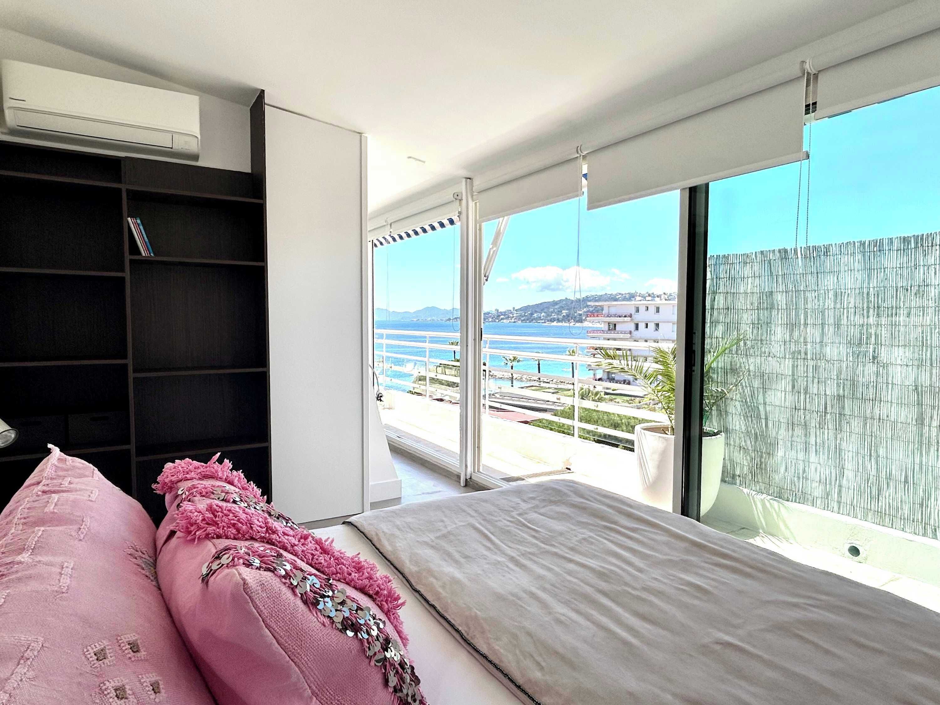 A bedroom with a large window, a bed, a wardrobe, and a view of the sea and buildings.