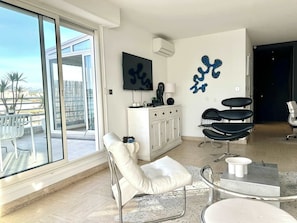A modern living room with a large sliding glass door, a flat-screen TV, a white cabinet, a coffee table, and a seating area with a white chair and a black chair. - Riviera rooftop - front de mer avec terrasse (Antibes)