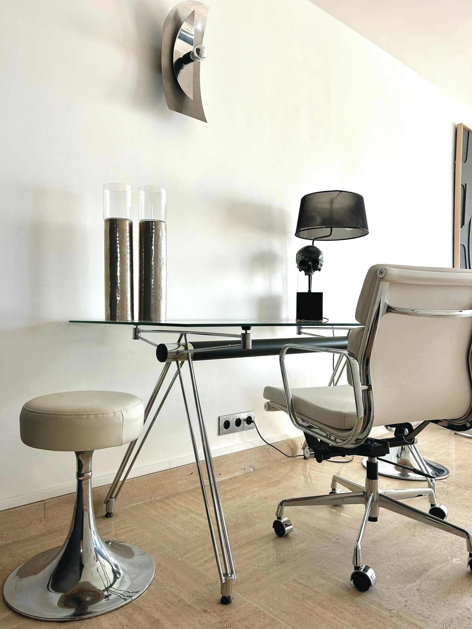 A modern office chair, a glass table with metal legs, and a wall-mounted lamp.