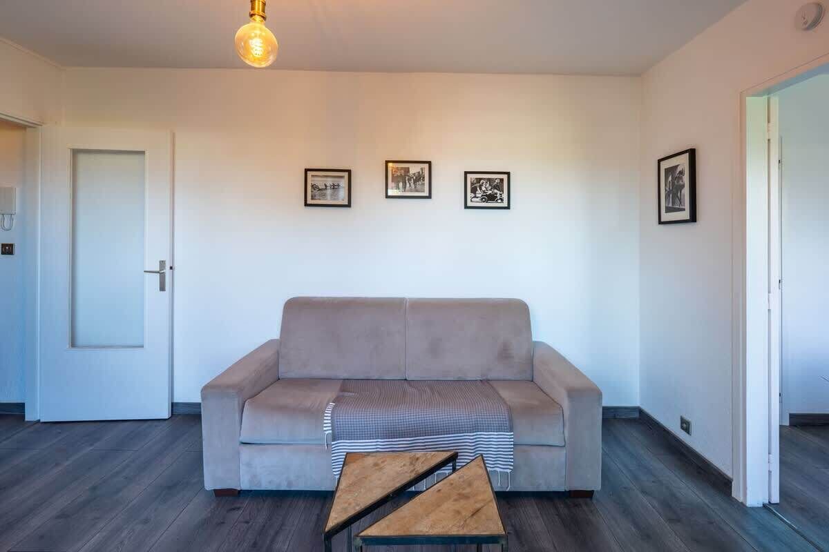 Sala de estar em tons neutros, com um sofá, uma pequena mesa de centro de madeira e um lustre. Há três quadros na parede.