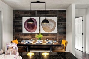 Un comedor con pared de madera oscura, dos lámparas colgantes, una mesa preparada para cuatro comensales y una puerta a la derecha.