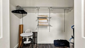 Una habitación compacta con una pared blanca, una pequeña mesa blanca, una silla negra, un estante metálico con toallas dobladas y un bolso negro en el suelo.