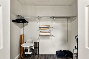 Ein kleines Zimmer mit weißer Wand, einem kleinen weißen Tisch, einem schwarzen Stuhl, einer Metallregaleinheit mit gefalteten Handtüchern und einer schwarzen Tasche auf dem Boden.