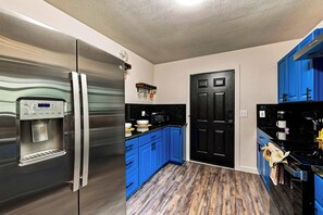 Una cocina con un refrigerador de acero inoxidable, armarios azules, una puerta negra y piso de madera.