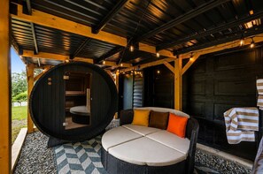 A covered outdoor seating area with a circular sauna, a wicker sofa with cushions, and a patterned floor. - Rolling Hills Retreat | Hot Tub • Fire Pit • Sauna (Pigeon Forge)