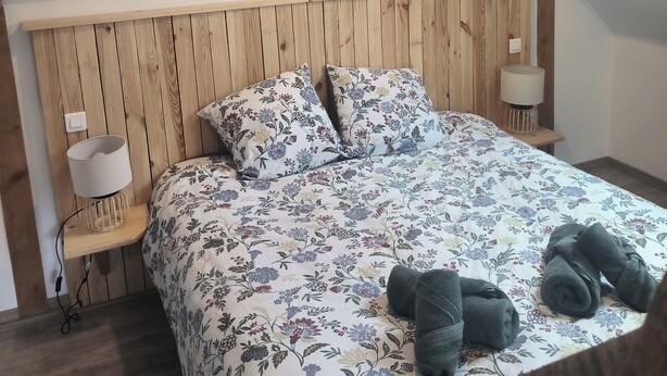 Een slaapkamer met een schuine kap, houten hoofdbord, bloemenbedrukking, een bedlamp en een klein raam.