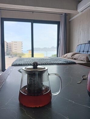 A room with a bed, a window with a view of buildings and a body of water, and a table with a glass teapot.