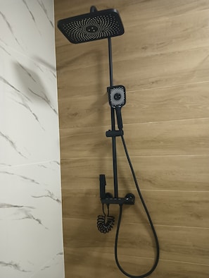 A black shower system with a handheld showerhead and a handheld scrubber against a wooden wall.