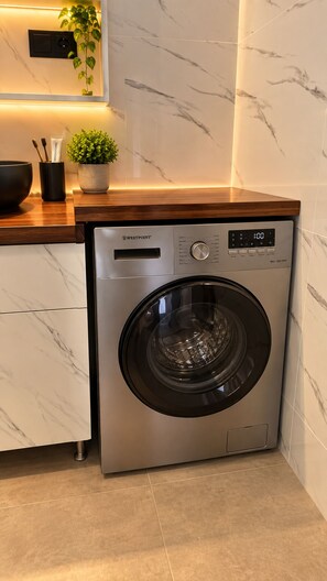 A compact, stainless steel front-loading washing machine with a digital display, situated in a modern kitchen with marble countertops and a wooden countertop. - Панорамный вид на океан, бухту, город. Спокойный район. Песчаный каньон. (Lazareto)