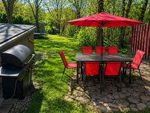 En udendørs terrasse med grill, røde stole og et bord under en rød parasol.