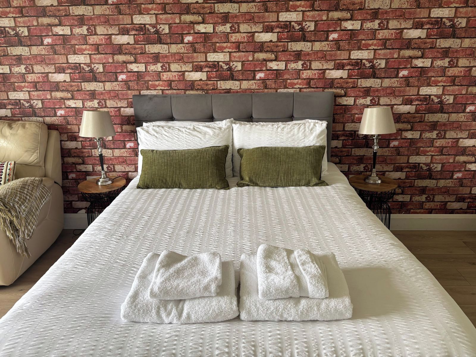 A neatly made bed with a white textured bedspread, two green pillows, and a pair of folded white towels.