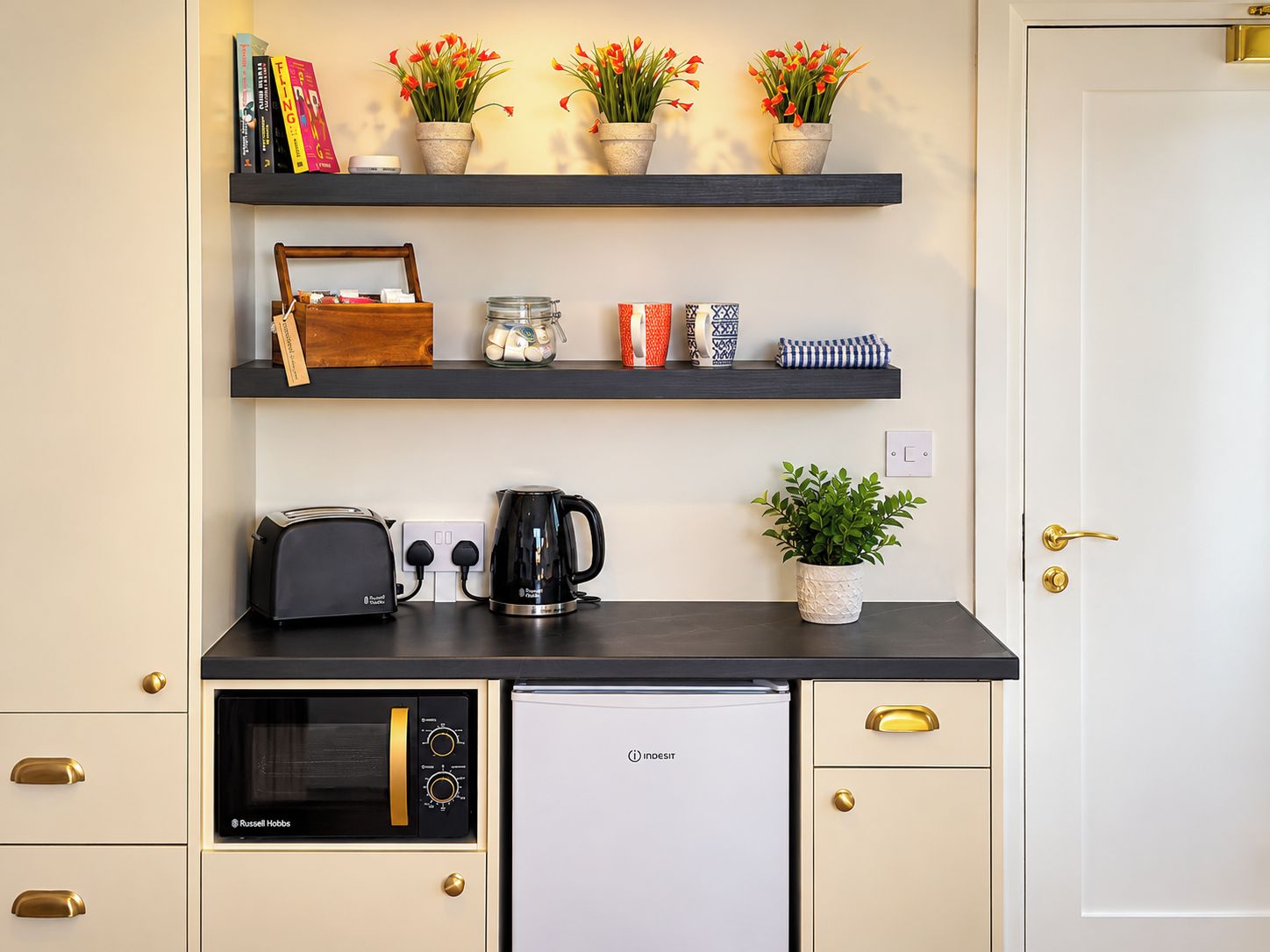 A kitchenette with a microwave, toaster, and coffee maker. There are three shelves above with books, a jar of tea, and decorative items.