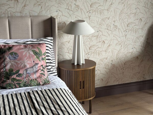 A bed with striped bedding, a round wooden nightstand with a lamp, and a wall with floral wallpaper.