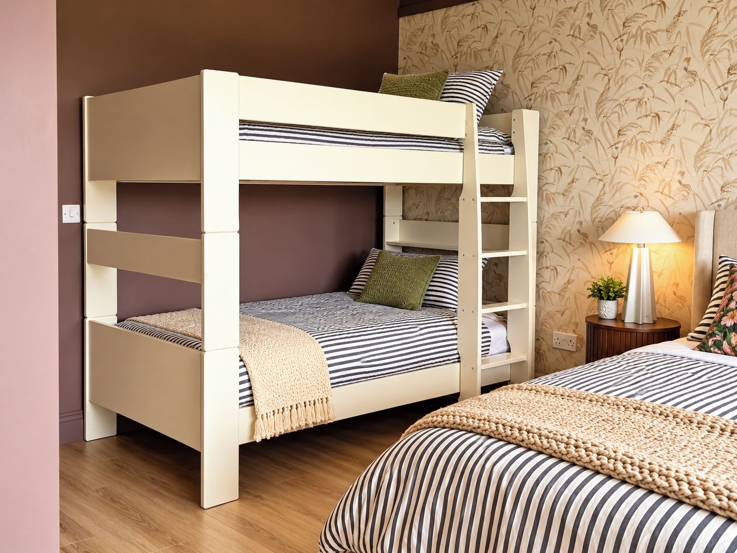 A bunk bed with a striped mattress, a small bedside table with a lamp, and a wall with a floral wallpaper pattern.