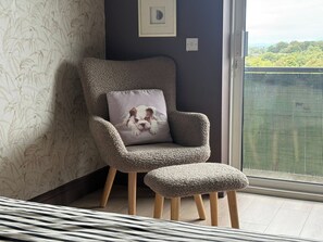 A corner seating area with a plush armchair and ottoman, a cushion with a dog print, a patterned wallpaper, and a view of greenery through a window.