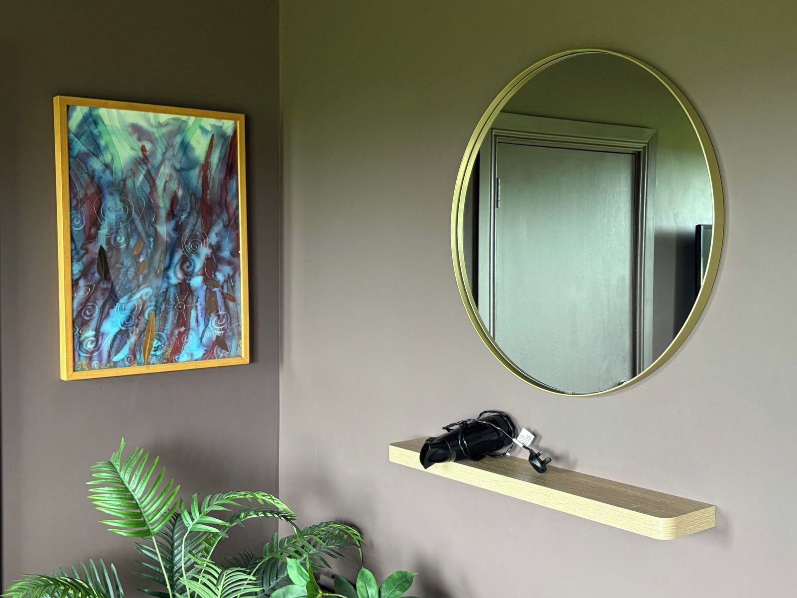 A room with a framed abstract painting, a mirror, a shelf with a black bag, and a potted plant.