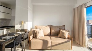 A modern living room with a sofa, a dining table with chairs, a kitchen area, and a large window with curtains. - Studio in Condo w/ Pool in Setor Bueno Bli2903 (Goiânia)
